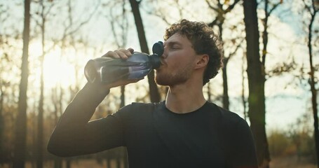 Confident male athlete with curly hair drinks water from a special gray sports bottle while resting after a run in the autumn forest on a sunny day. A man in a black sports uniform drinks water and