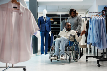 African american man in wheelchair choosing apparel while shopping with girlfriend in clothing...