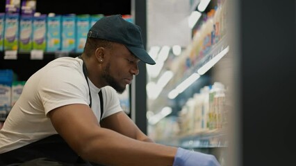Close-up shot of a man with Black skin in a white T-shirt and a black apron laying out products in a dairy display case in a large supermarket