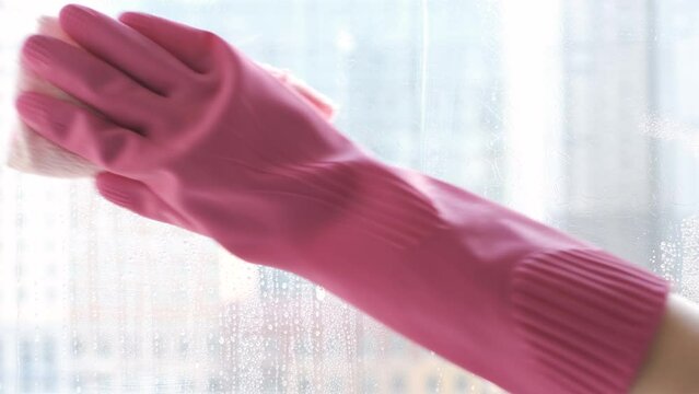 Female hand in pink gloves washes the window with rag and sprays detergent, spring cleaning concept.