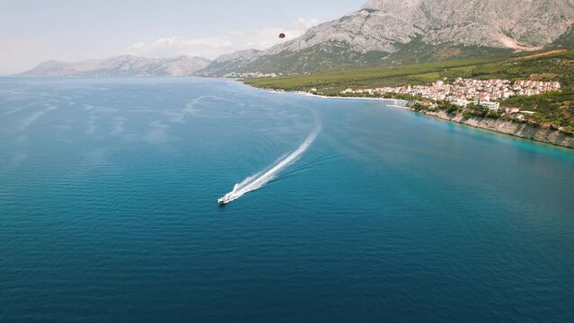 Speedboat carves a frothy trail in the calm Adriatic Sea. Above, a red parasail floats under a clear sky.