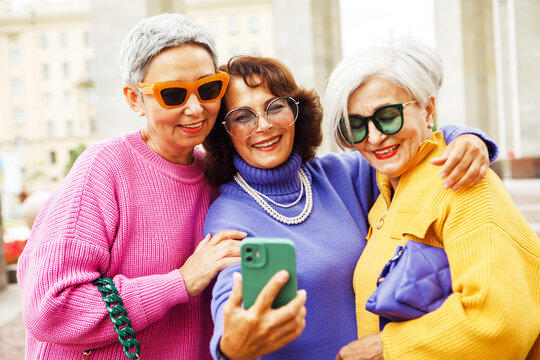 Three cheerful elderly friends in bright sweaters take a selfie on a smartphone and laugh, having a great time.
