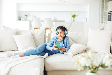 Cute child girl with pigtails with headphones and mobile phone on sofa indoors.