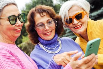 Three cheerful elderly friends in bright sweaters take a selfie on a smartphone and laugh, having a...