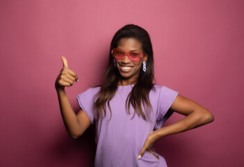 Funny black woman wearing liliac shirt and sunglasses showing ok sign over pink background.