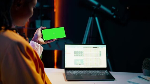African american girl holds phone with isolated greenscreen at home, studying economics and finances at university. Young woman uses blank mockup template while she works on spreadsheets. Camera B.
