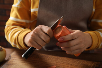 Woman peeling fresh carrot with knife at wooden table indoors, closeup