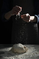 Making bread. Woman sprinkling flour over dough at table on dark background, closeup
