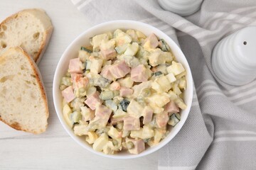 Tasty Olivier salad with boiled sausage in bowl and bread on white wooden table, flat lay
