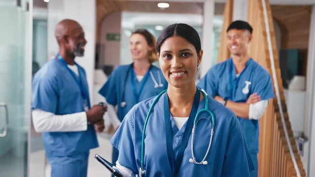 Close up portrait of smiling female doctor or nurse with digital tablet wearing scrubs standing in hospital corridor with multi-cultural team of colleagues in background - shot in slow motion