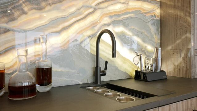 Luxury bar counter in warm colors with wall decorated with marble patterned tile, wooden cabinets for storing dishes, decanters for alcohol and sink