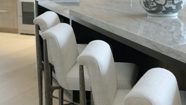 Row of white bar stools with gray legs standing next to reflective marble dining surface with patterned fruit bowl in open kitchen with oven and light floor