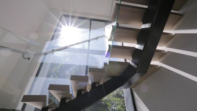 Bottom view of elegant staircase with glass railings inside building illuminated by natural light coming through panoramic window. Wooden steps constructed with dark frames