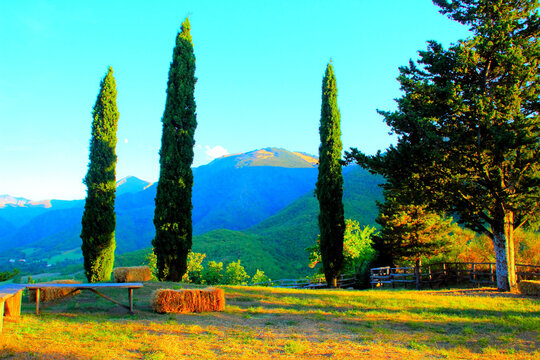Warm view from Fiastra at Giardino delle cince with some benches and a parallelepiped bale of hay used as seating, a meadow with tall green trees in the foreground and the huge Sibillini in the back