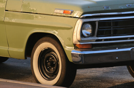 A partial view of the front end of a classic, vintage green Ford F-100 truck with a white wall tire, chrome molding, and bumper