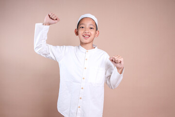 Young Asian Muslim boy wear cap clenched fist celebrating victory over beige background