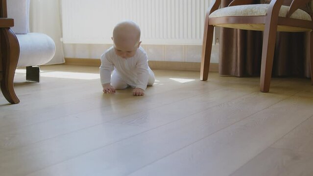 Curious baby crawl on wood floor, explore sunlit room, white onesie, home interior. Cute Infant on all fours in bright room, elegant home area setting, soft natural lighting. Child navigate floor.