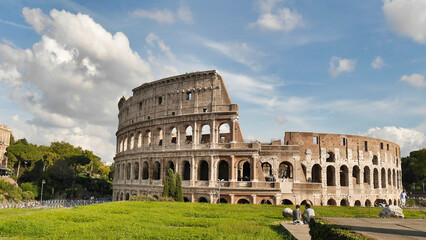 The Colosseum stands under a sunny sky, surrounded by greenery and ruins, showcasing its iconic...