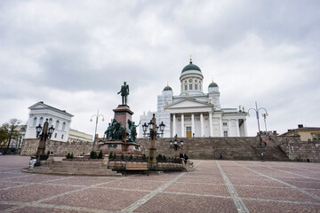 A wide square with people, a central statue, and yellow buildings, under an overcast sky.