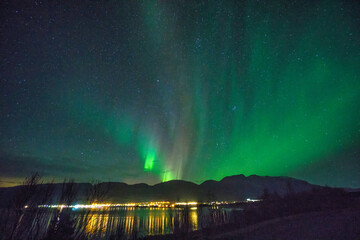 The Northern Lights (Aurora Borealis) spread across the night sky above a mountain range with city...