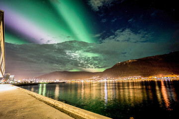 The Northern Lights (Aurora Borealis) spread across the night sky above a mountain range with city...