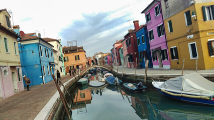 Colorful houses reflect on the canal in Burano, Venice, with boats and a bridge, showing the...