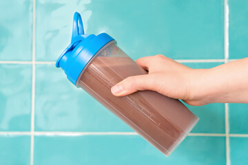 Female hand holds shaker with protein shake against background of mint tiles.