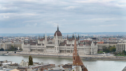 The Hungarian Parliament Building beside the Danube River, under a cloudy sky, with city buildings in the foreground.