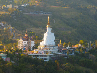 Aerial view of a majestic white Buddha statue atop a hill with a scenic sunset backdrop, overlooking a bustling town below.