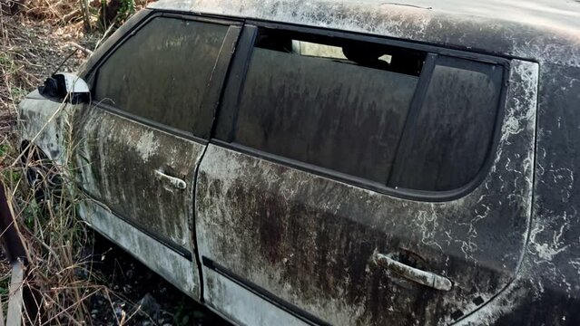 Abandoned white car with damaged paint and broken windows overgrown with vegetation depicting neglect and decay old accident scrap car