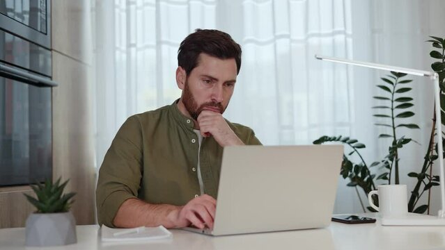 Tired man entering data into laptop and rubbing temple while taking notes during busy day in home workspace. Thoughtful boss analysing results of coworkers and preparing remarks for online meeting.