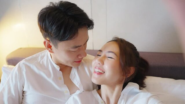 Asia people young adult man woman relax kiss hug smile on home bed looking at video camera holding mobile enjoy take photo record vlog. Joy happy asian lovers wife husband just married sweet newlywed.