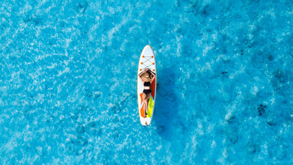 Aerial view of a beautiful young woman in black swimsuit lying on Paddle Board  on a blue sea with...