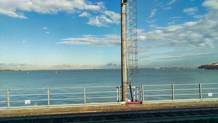 A coastal landscape is viewed through a train window, showing a calm sea and distant buildings...