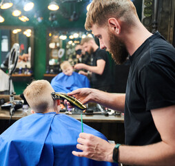 Hairdresser shaving boy's head. Competent barber using electric shaver at barbershop. Little client...