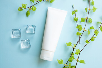 White Cream tube, young small leaves of birch tree, ice cubes on blue. Cosmetic skincare product blank plastic package. Bottle of moisturizer, sunscreen lotion, balsam, toothpaste mockup, flat lay. - obrazy, fototapety, plakaty