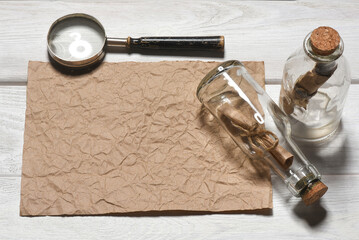 Message scroll letter in the bottle, magnifying glass and blank crumpled blank paper sheet with copy space on the white wooden desk table flat lay background. Ancient map concept.