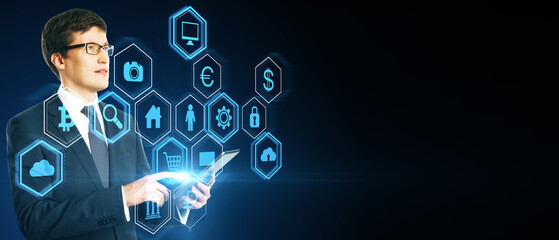 Attractive young european man using tablet with abstract hexagonal pattern hologram with various business icons on wide dark background with mock up place. Digital dashboard and data concept.