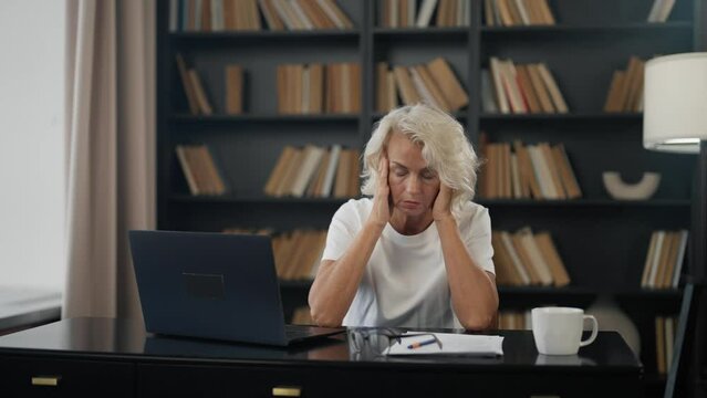 Mature woman suffer headache at work. Senior female has migraine sitting at table at home office rubbing temples. Tired exhausted woman feeling pain fatigue. Menopause symptoms chronic stress nervous.