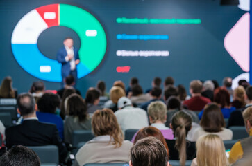 Focused audience at a business conference listening to a presenter with a pie chart on screen.