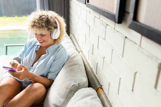 Woman relaxes on a couch, listening to music with white headphones and holding a smartphone. The indoor setting shows a comfortable and modern lifestyle, with a touch of technology and leisure.