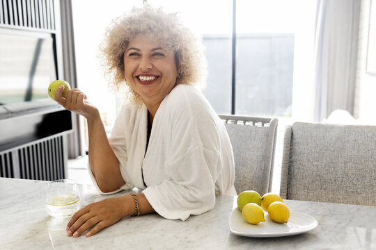 Cheerful curly-haired mature woman holding an apple sitting in a sunlit kitchen. A moment of happiness and healthy lifestyle captured with a fresh and cozy home atmosphere.