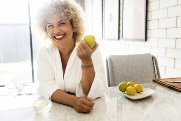 Bright and lively image capturing a joyful woman with curly hair, smiling while holding a green apple in a modern, sunlit kitchen setting. A sense of health and positivity emanates from the scene. - 741310784