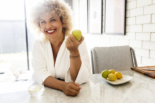 Bright and lively image capturing a joyful woman with curly hair, smiling while holding a green apple in a modern, sunlit kitchen setting. A sense of health and positivity emanates from the scene.