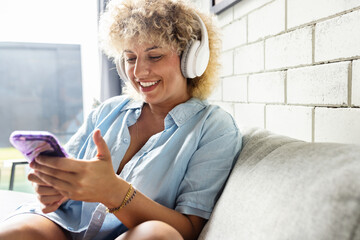 A cheerful young woman with curly hair listens to music on her white headphones while holding a smartphone, enjoying a relaxing moment on a couch by a bright window. - 741311390