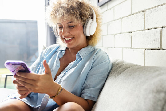 A cheerful young woman with curly hair listens to music on her white headphones while holding a smartphone, enjoying a relaxing moment on a couch by a bright window.
