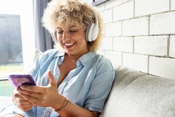 Woman Enjoying Music on Her Smartphone with Headphones. Female With Curly Hair Relaxes , Enjoying Podcast on her phone While Wearing White Wireless Headphones - 741311593