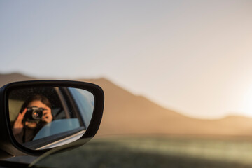 Chica sacándose un retrato en el reflejo del espejo retrovisor del coche durante las vacaciones, con una cámara de fotos - obrazy, fototapety, plakaty