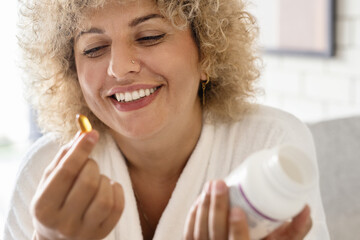 Morning Wellness Routine. Happy Woman in White Robe Taking Vitamin Capsule at Home. She holding a bottle, considering her daily health routine in a bright, cozy home setting. - 741313520