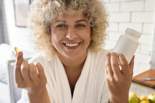 Close-up of a radiant woman in a white robe showing a nutritional supplement, promoting a routine of health and wellness at home with a smile.